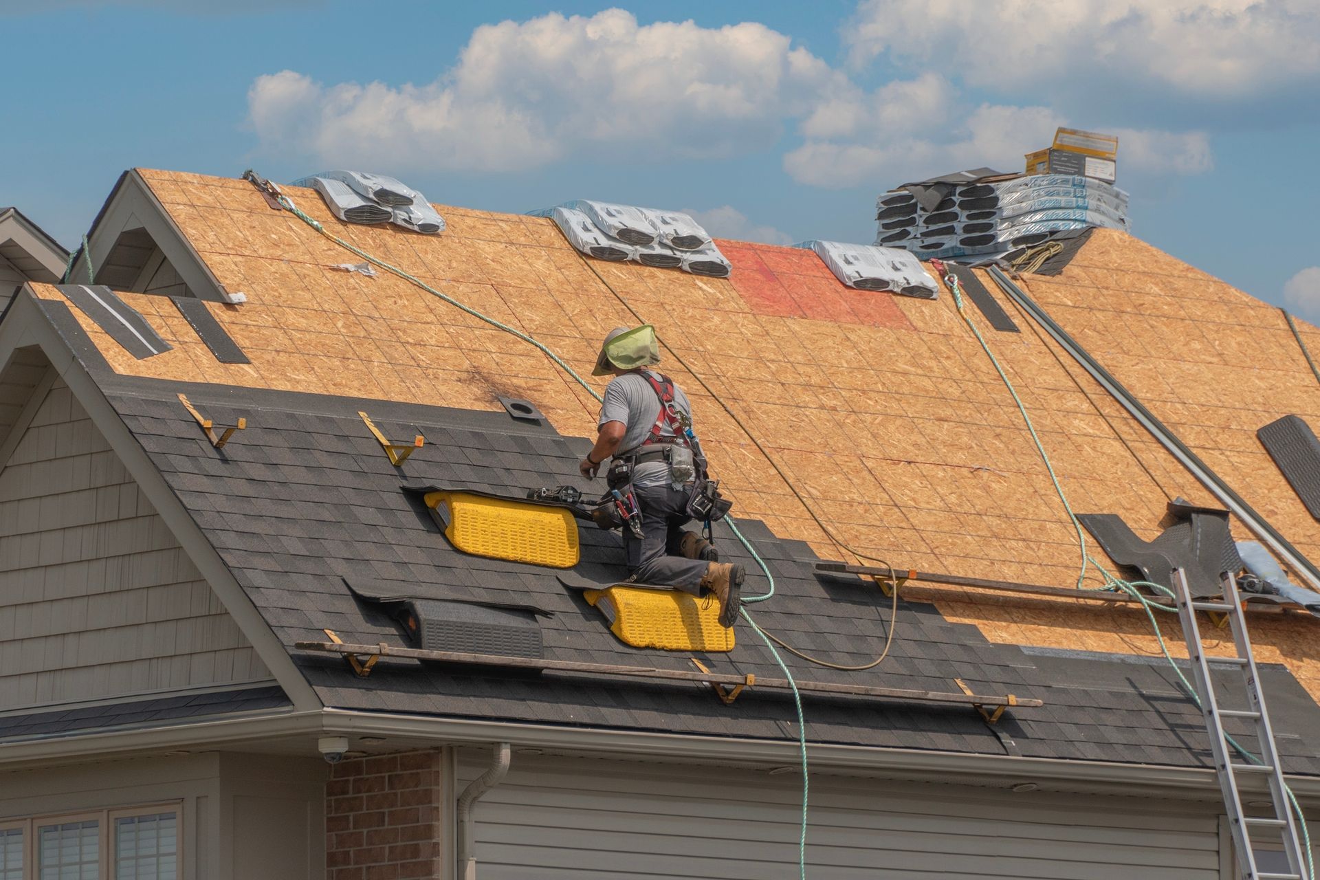 A construction worker wearing a safety harness stands on a residential roof installing new shingles.