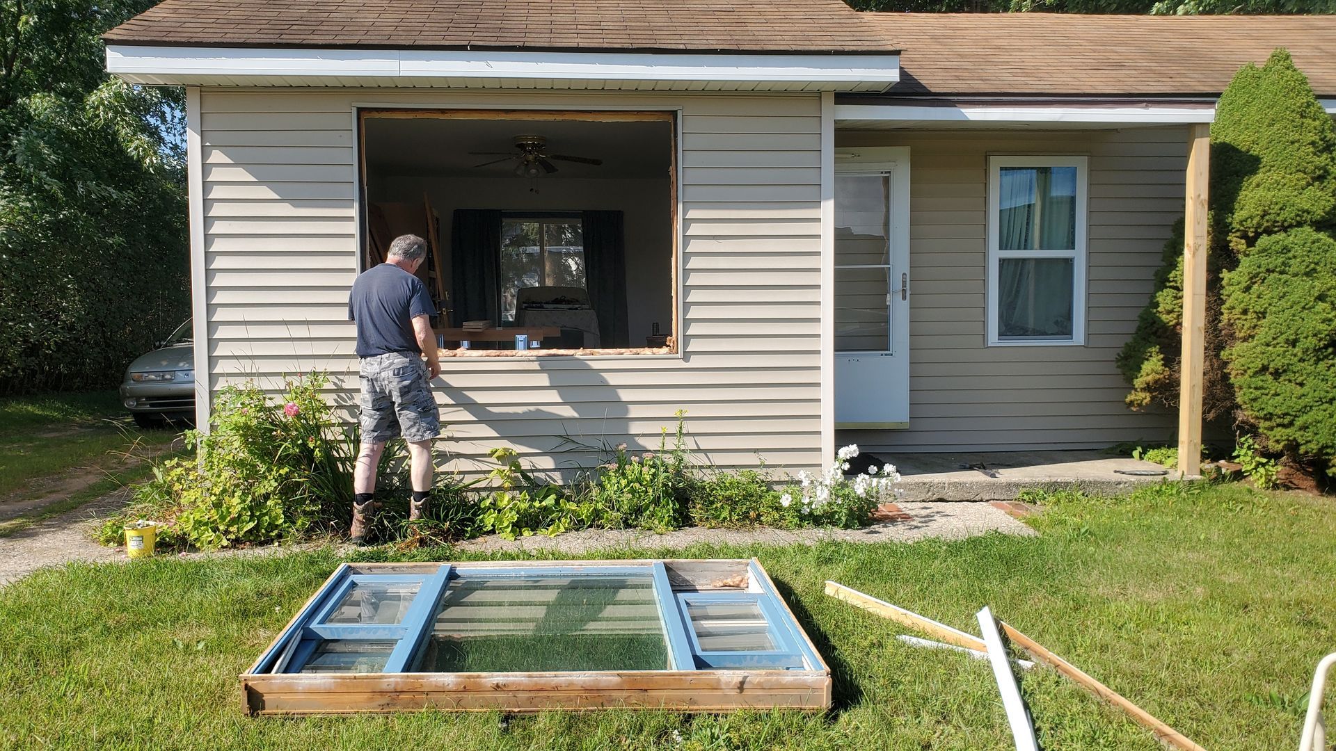 A person standing before a house with a large wall opening where a window unit lies on the grass in the foreground.