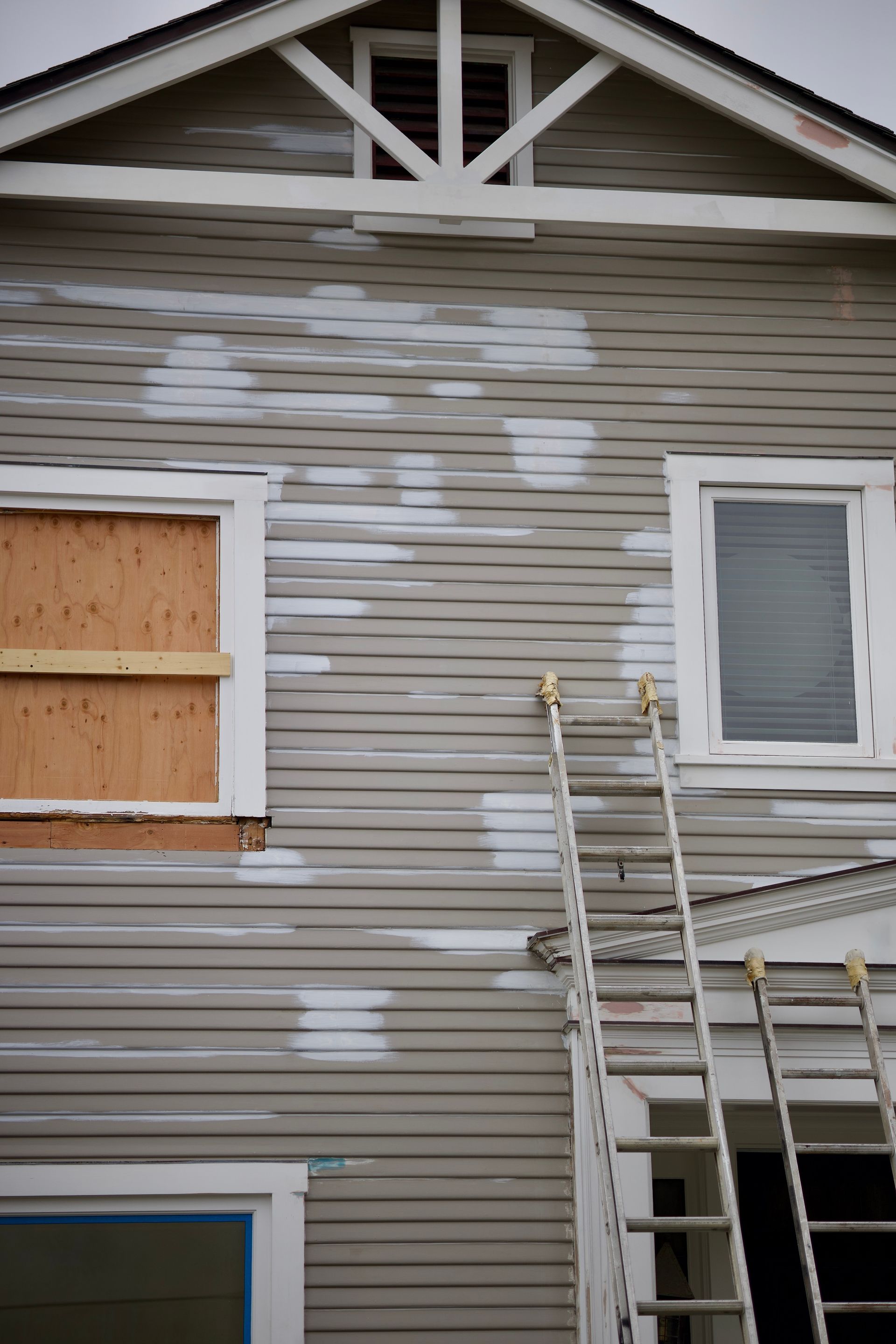 An exterior wall of a house with grey horizontal siding, white trim, a boarded window, and an extension ladder leaning up.