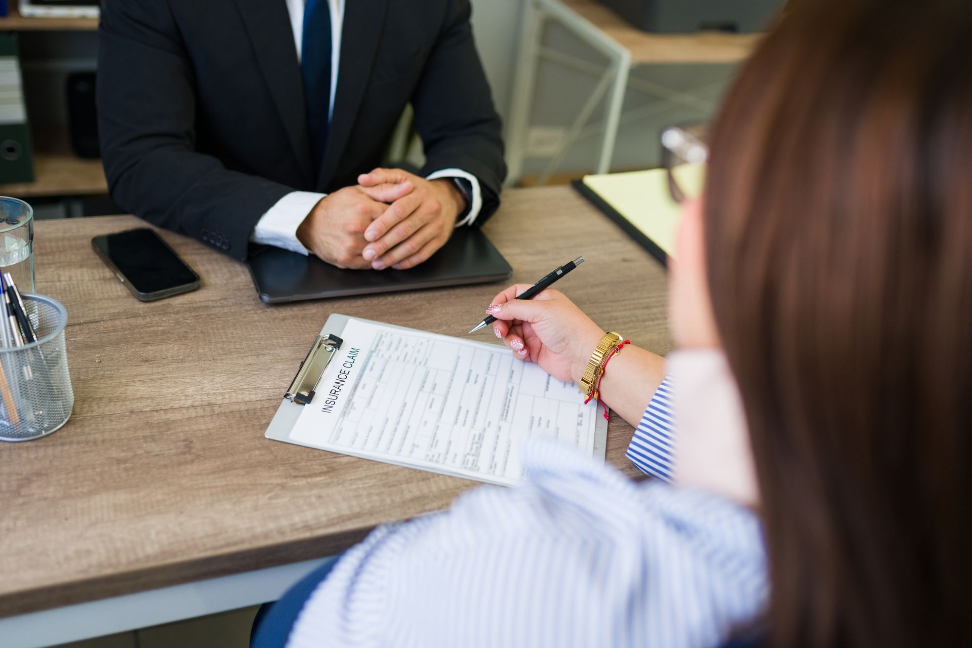 A person in a suit sits across from someone writing on a document at a wooden office desk.