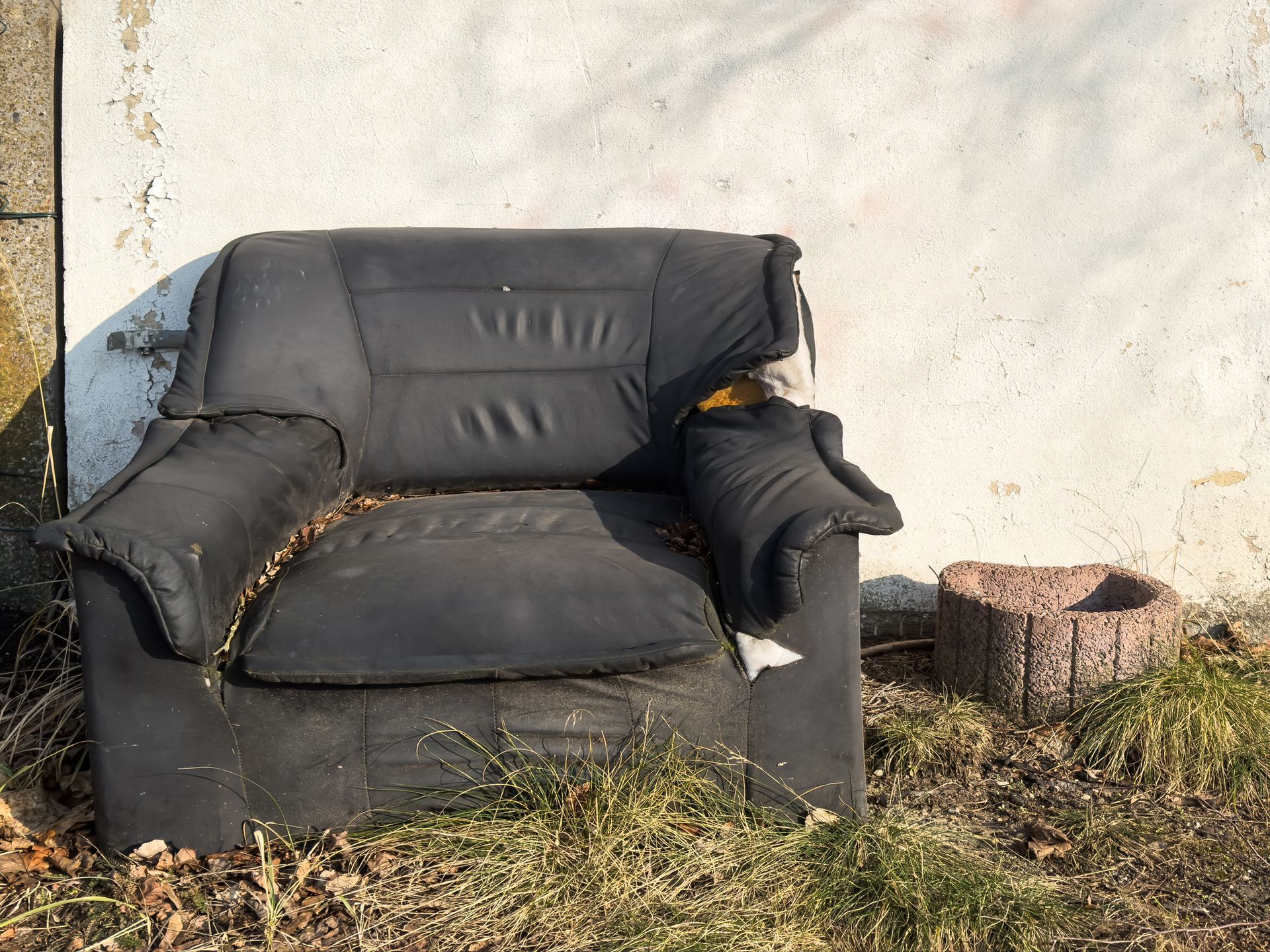 A worn black armchair sits outdoors on dry grass near a white wall.