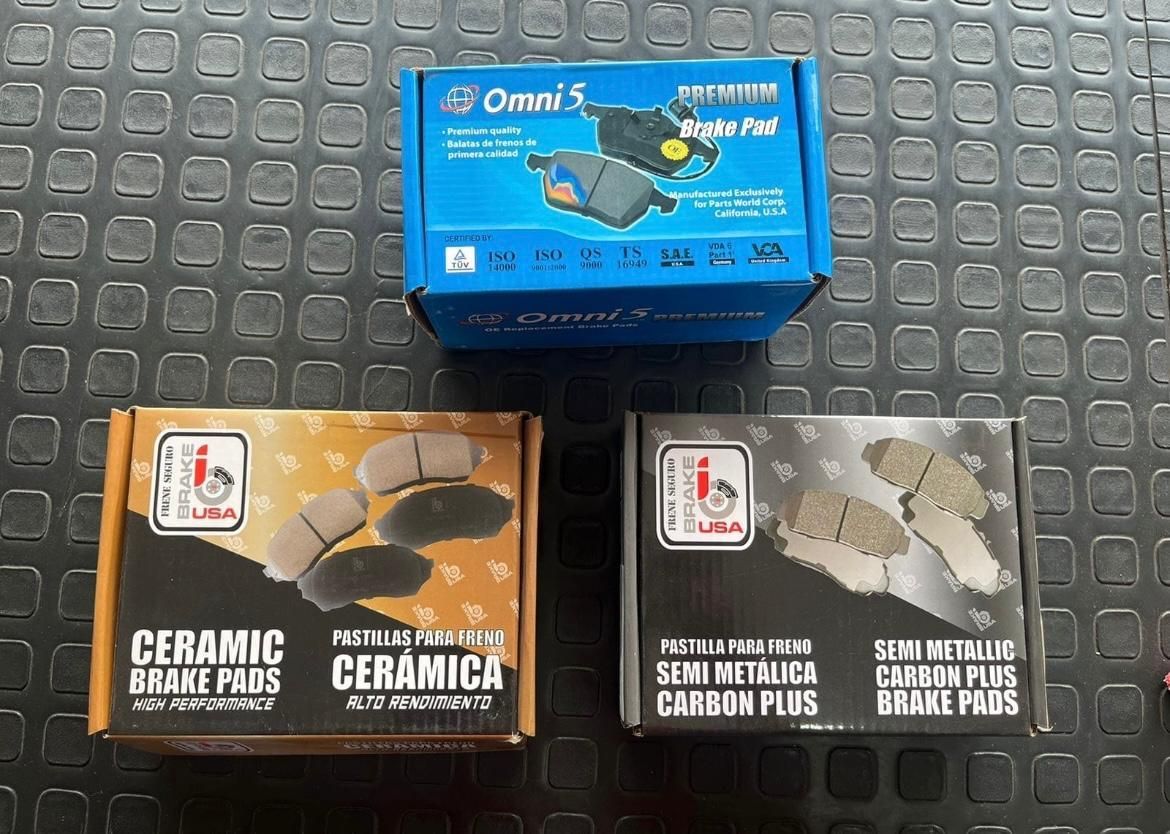 Tres cajas de pastillas de freno, azul, naranja y negra, sobre una superficie negra.