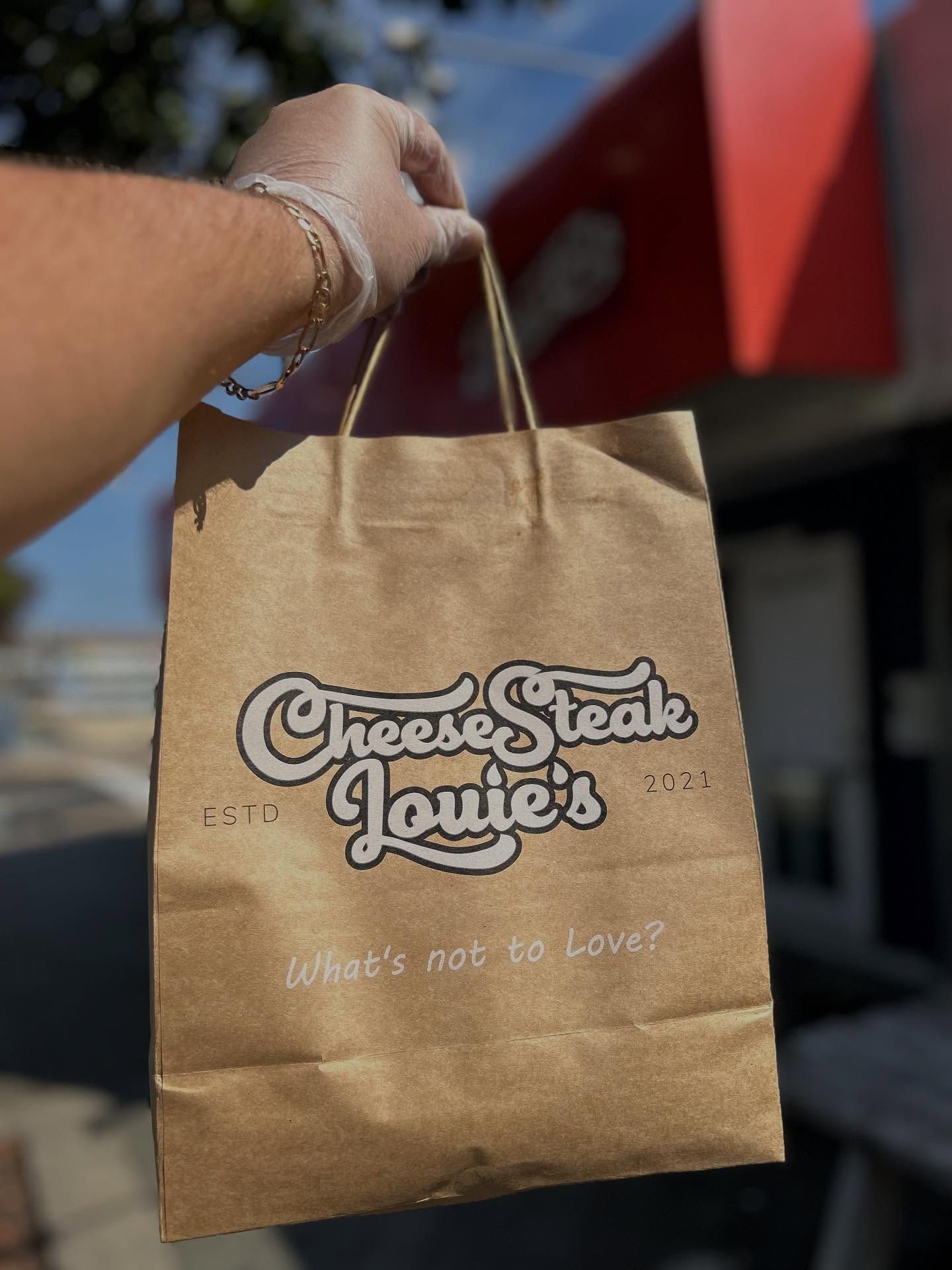 Hand holding a brown paper bag from "Cheesesteak Louie's". Bag has a logo and tagline: "What's not to love?"