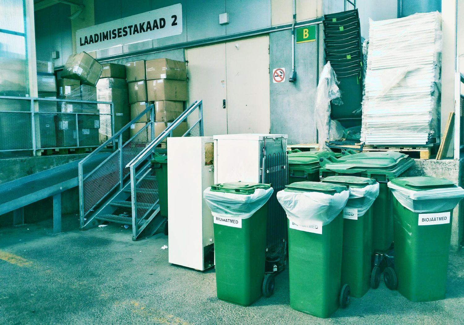 Green recycling bins in a loading dock area, boxes and waste stacked, building in background.