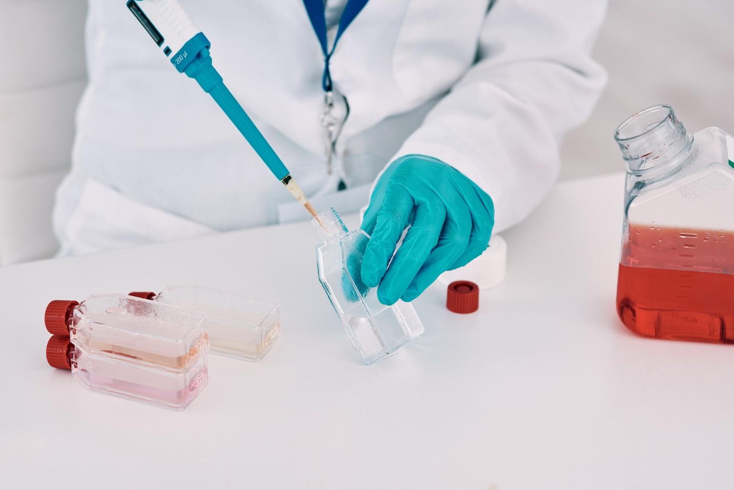Scientist in lab coat using a pipette to transfer liquid into a clear container, red-tinted liquid visible.