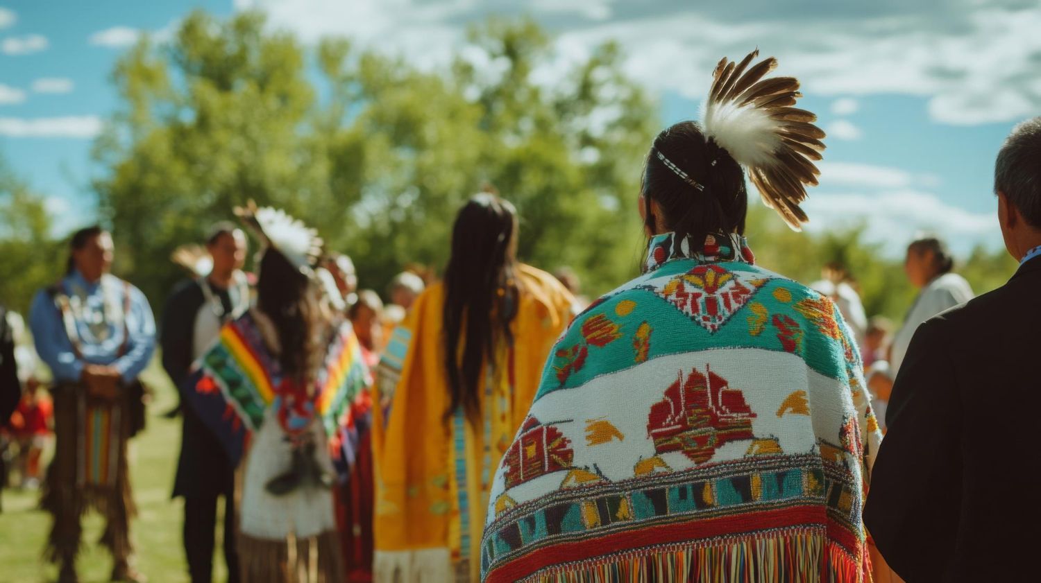 Indigenous people in traditional clothing at an outdoor gathering; colorful shawls and headdresses.