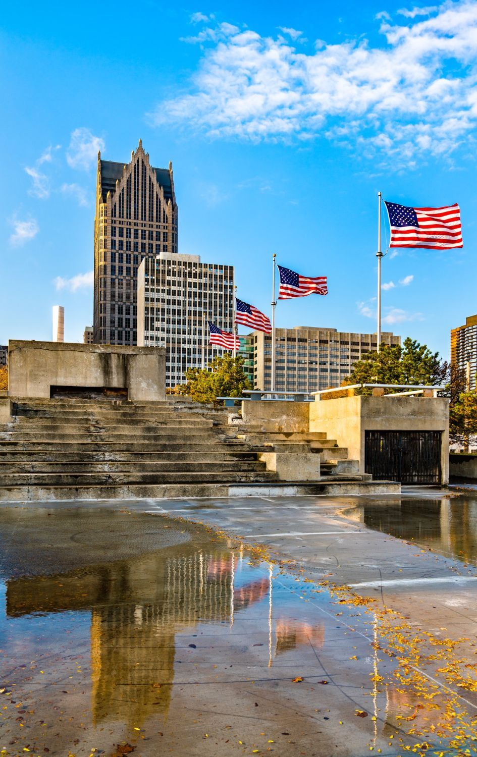 Detroit skyline reflected in a wet plaza; American flags wave.