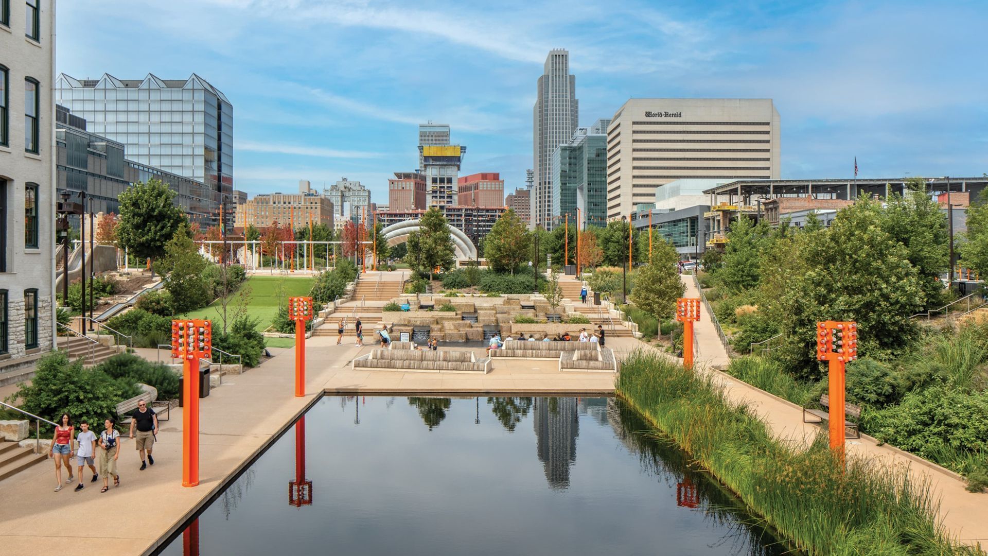 Urban park with reflecting pool, pathways, and modern buildings in the background. Orange accents and people walking.
