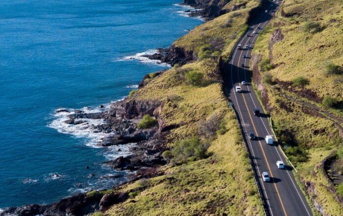 Coastal highway with cars traveling along a cliffside; blue ocean and green hills.