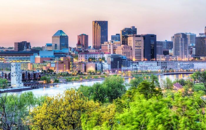 City skyline at dusk with buildings of various heights along a river, green trees in the foreground.
