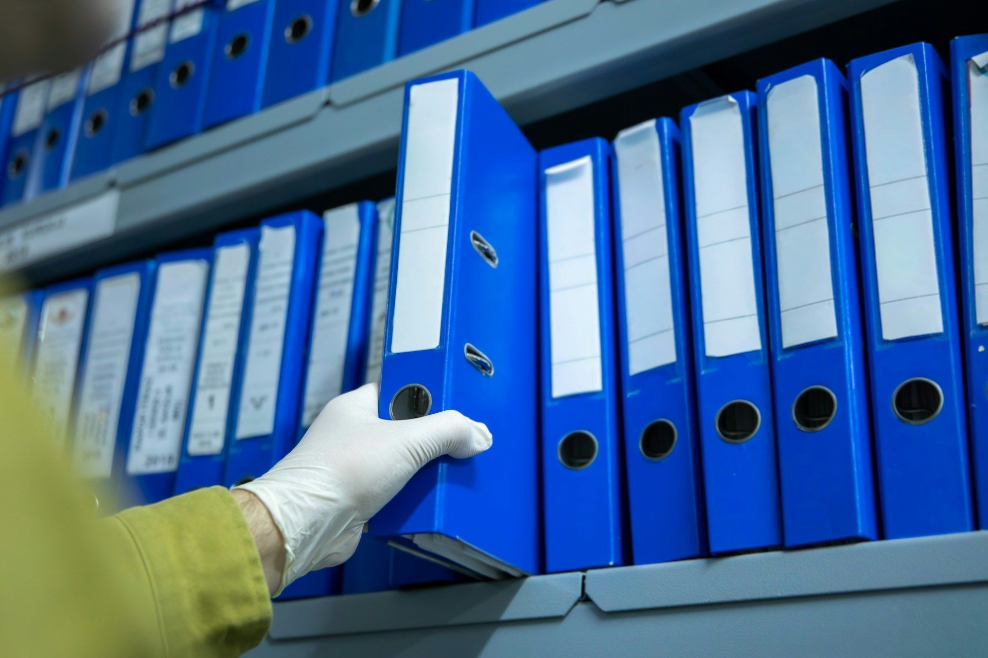 A person wearing a white glove pulls a blue binder from a shelf filled with similar organized records.
