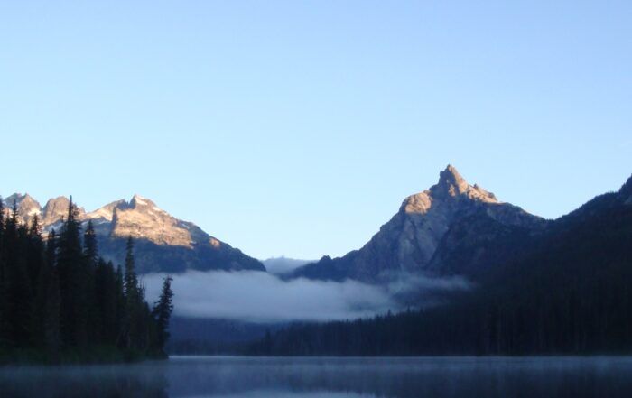 Mountains with sunlight peaks, fog over water. Dark trees frame the water's edge, clear sky above.