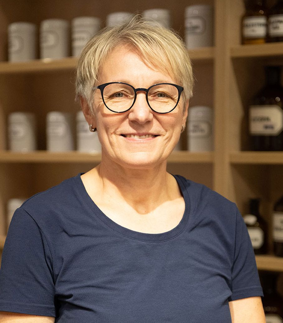 Eine Frau mit Brille und blauem Hemd lächelt vor einem Regal voller Flaschen.