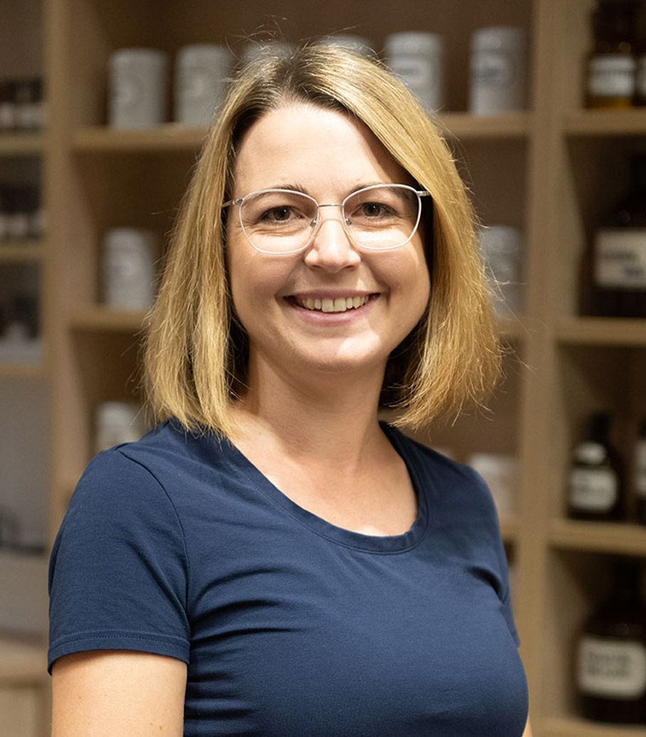 Eine Frau mit Brille und blauem Hemd lächelt vor einem Regal voller Flaschen.