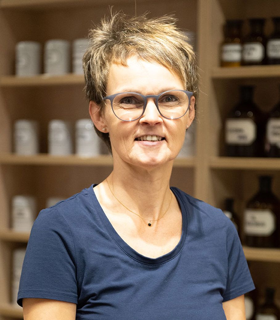 Eine Frau mit Brille und blauem Hemd steht vor einem Regal voller Flaschen.