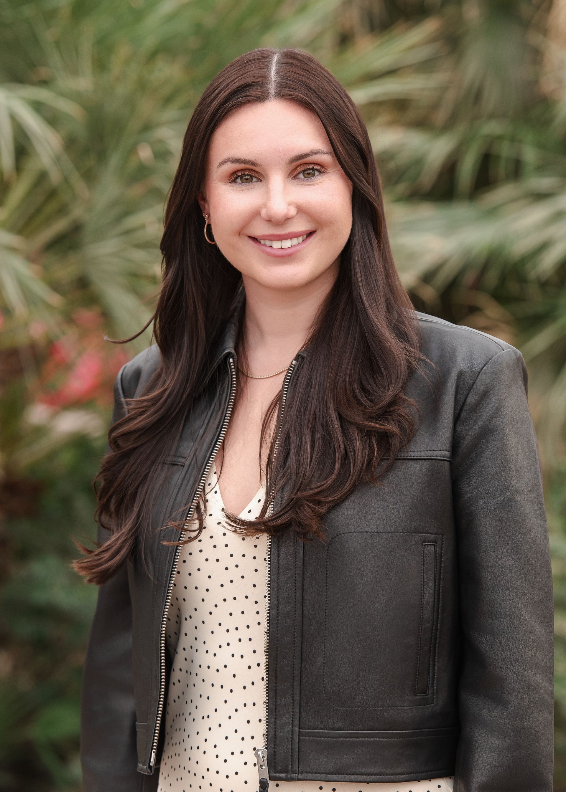 Woman with long brown hair, smiling, wearing a black jacket and dotted top, outdoors with greenery.