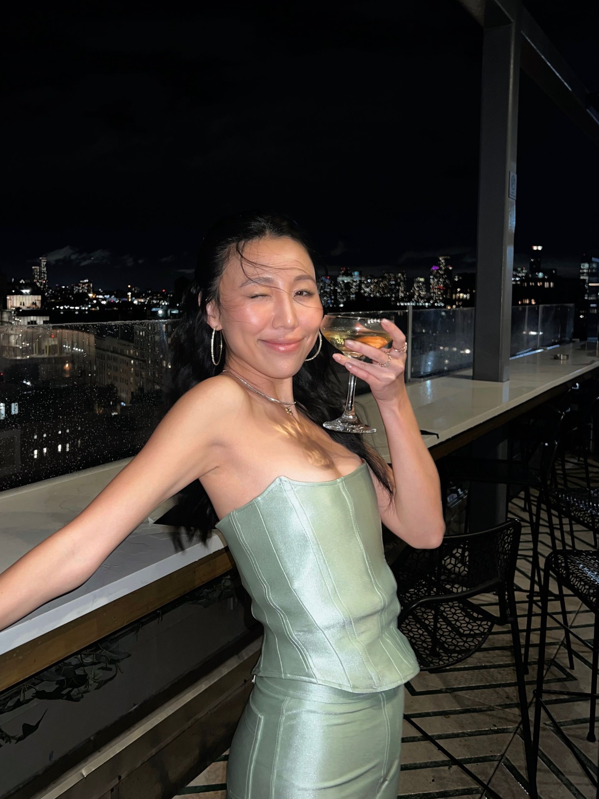 Woman in green corset dress on a balcony at night, holding a drink, smiling. City skyline in background.