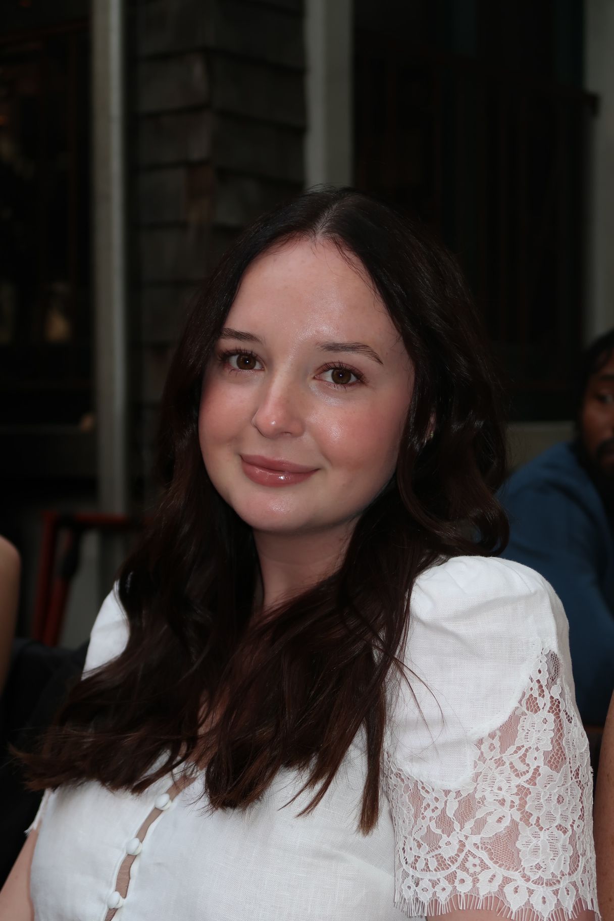 Woman with dark hair, wearing a white lace top, smiling, outdoors.