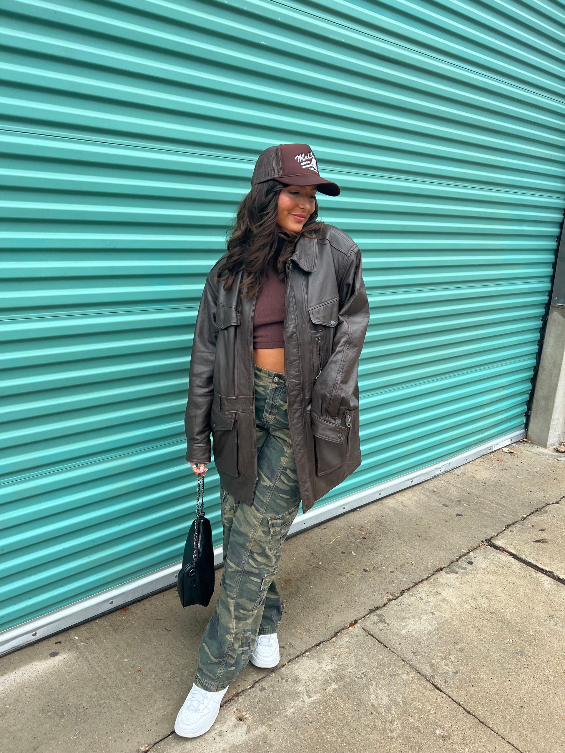 Woman in brown jacket, hat, and camo pants, smiling, standing by a teal garage door.