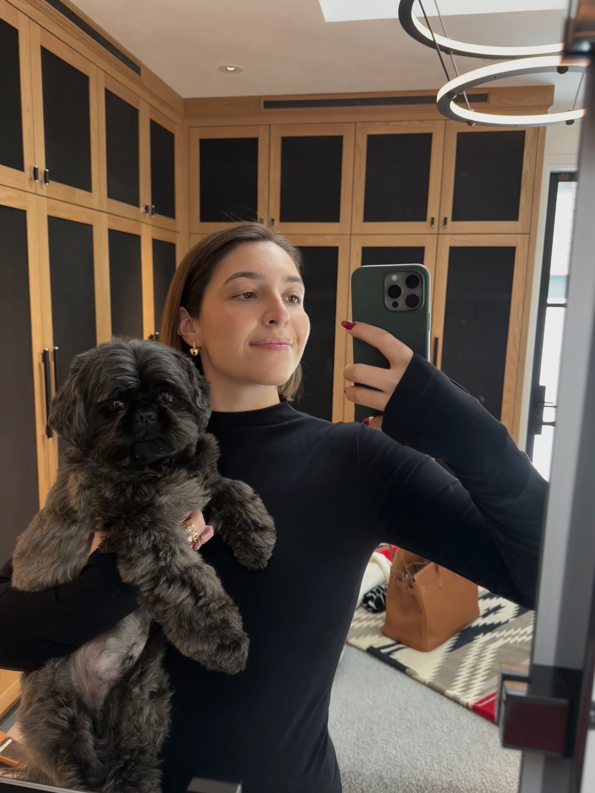 Woman holding small, dark-haired dog, taking a mirror selfie in a closet with dark wooden cabinets.