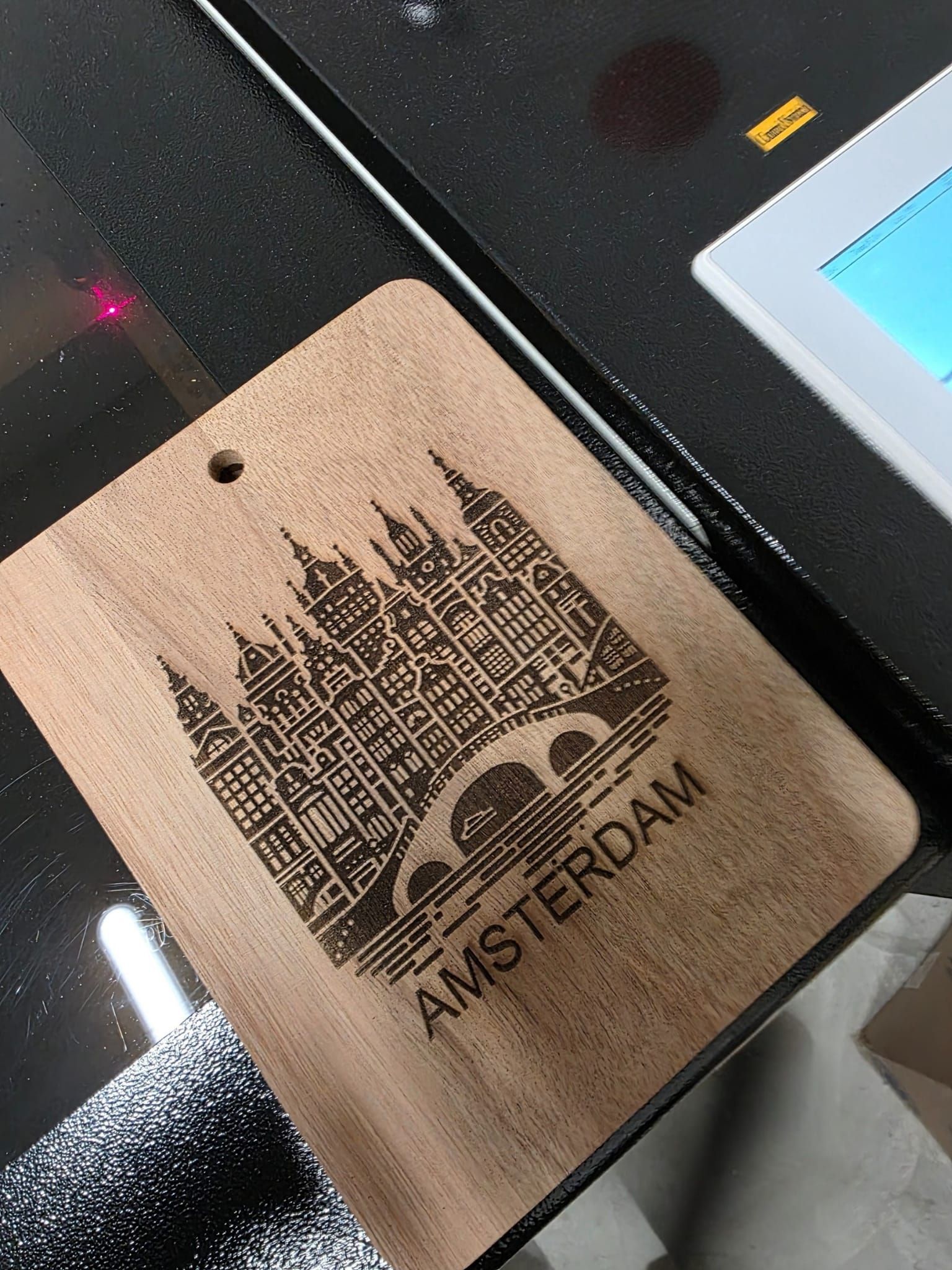 Wooden tag laser-etched with Amsterdam skyline and text, inside a laser engraving machine.