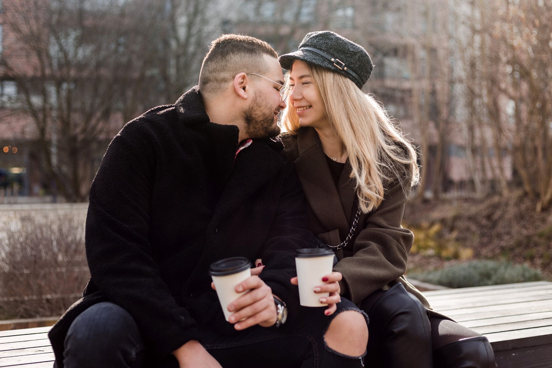 Two people in dark coats sit on a bench, smiling and sharing kisses while holding takeaway drinks.