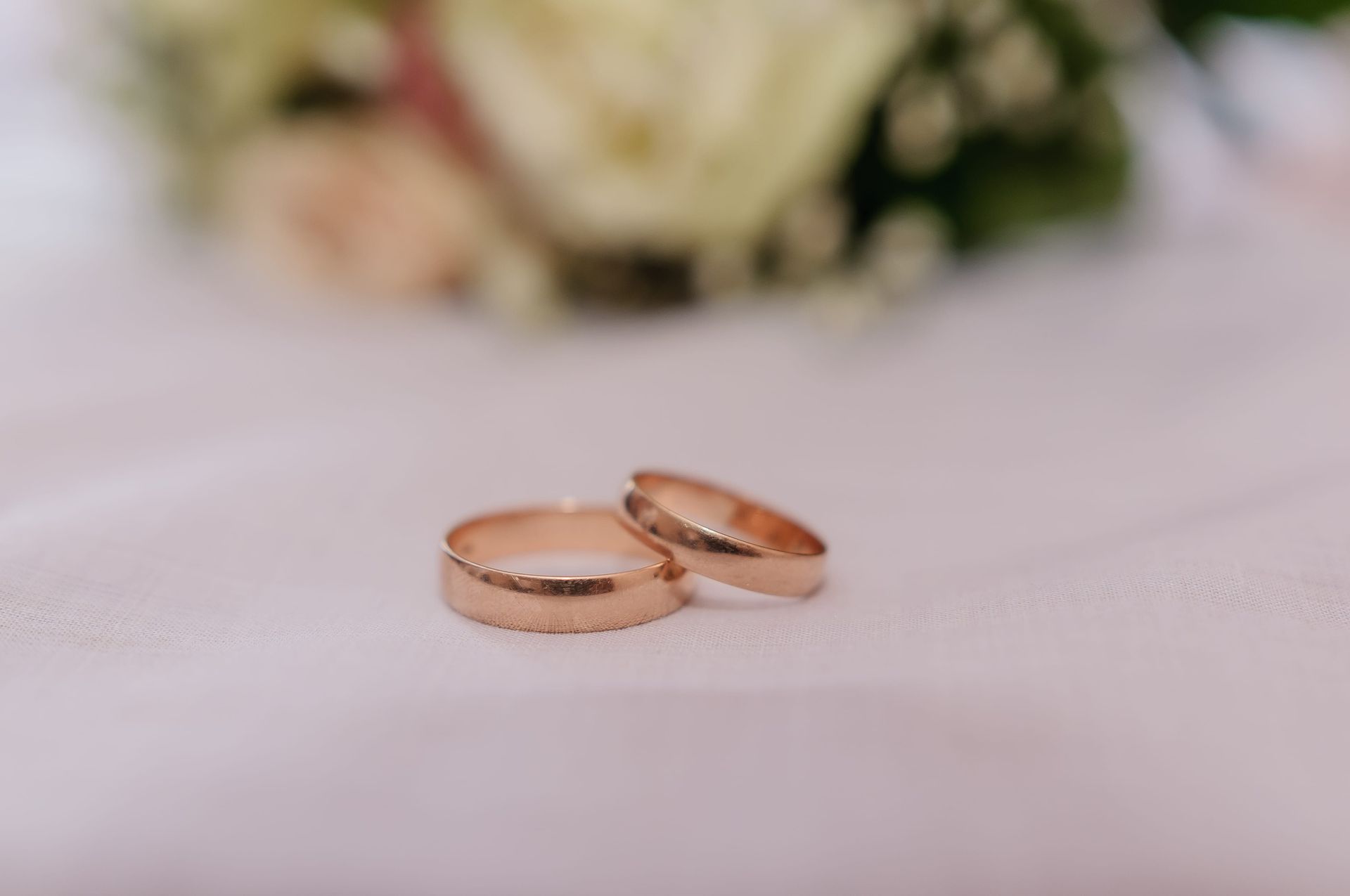 Two rose gold wedding rings on a soft white surface with blurred flowers in the background