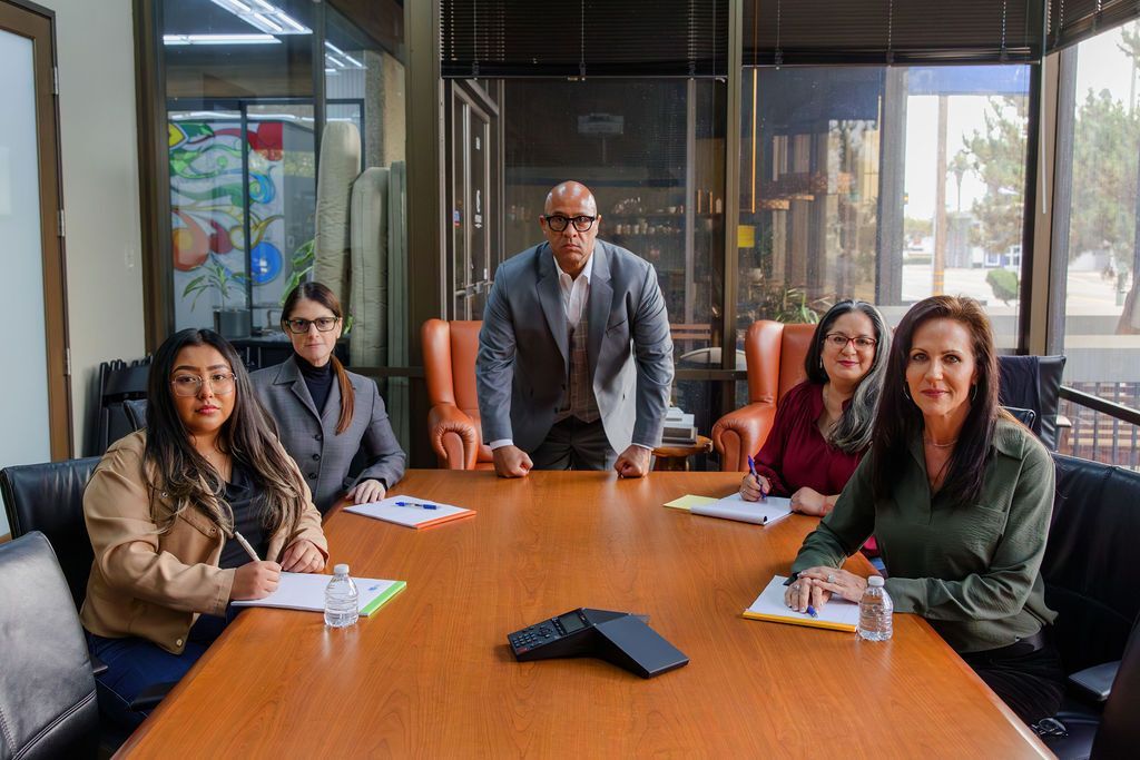 Five professionals in business attire sit and stand around a wooden conference table in an office, reviewing documents.