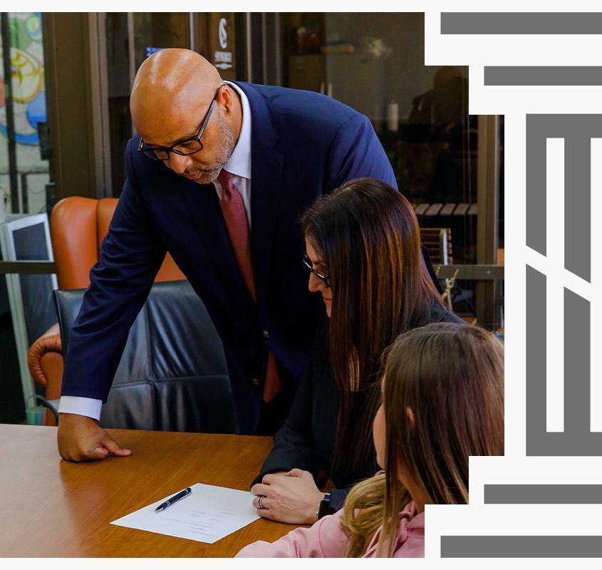 Three people in professional attire gather around a wooden desk, reviewing a document together in an office.