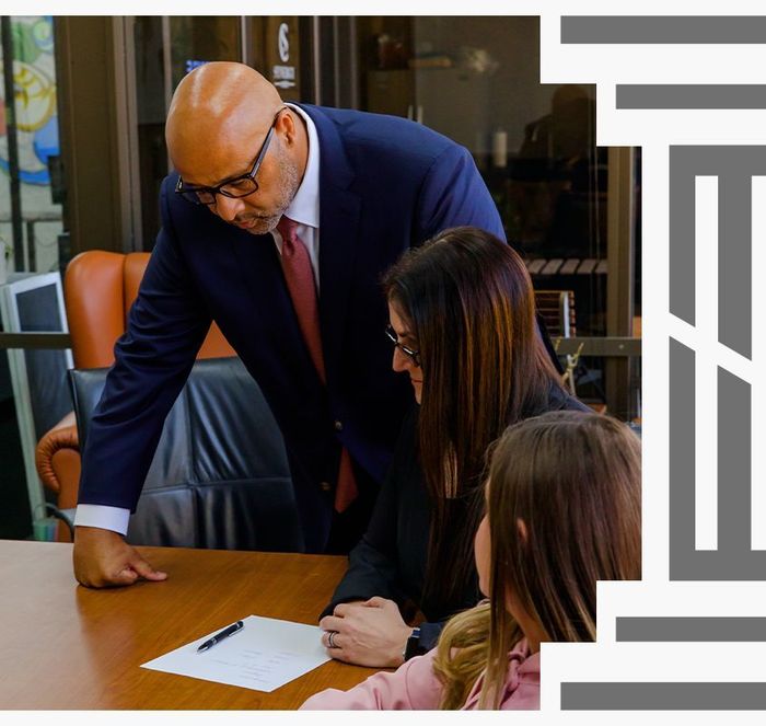 Three people in professional attire gather around a wooden desk, reviewing a document together in an office.