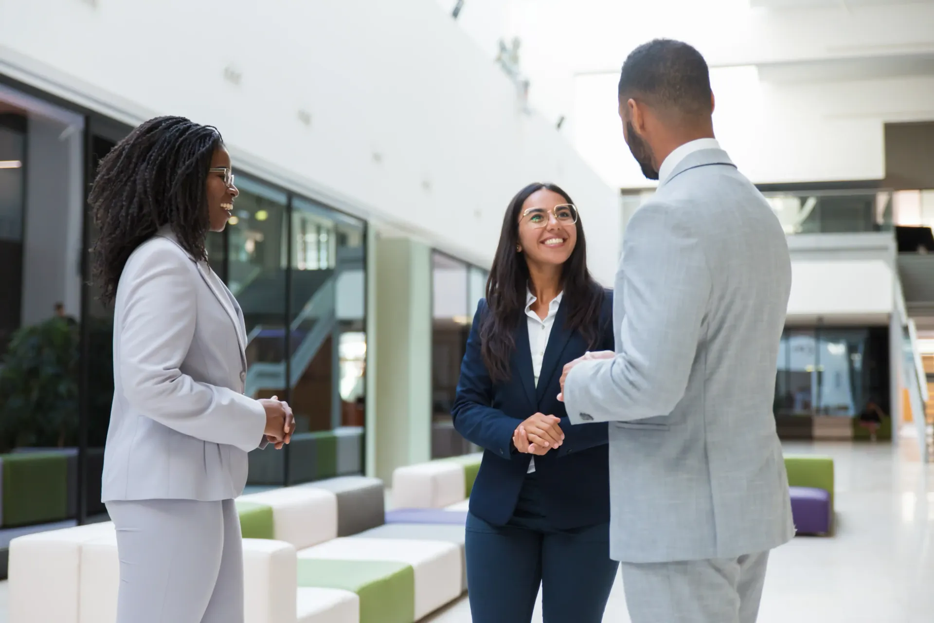 Three professionals in business attire converse while standing in a bright, modern office atrium.