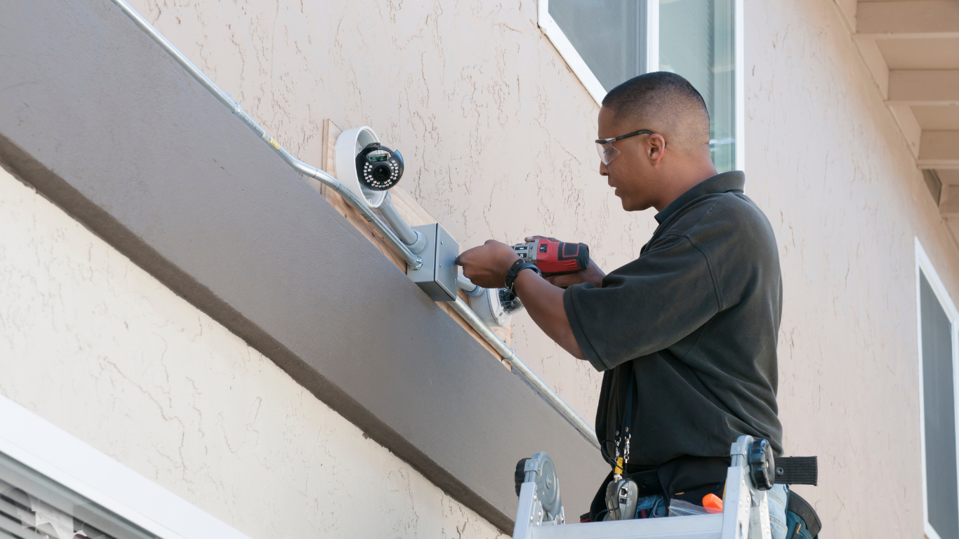 A man is working on a security camera on the side of a building.