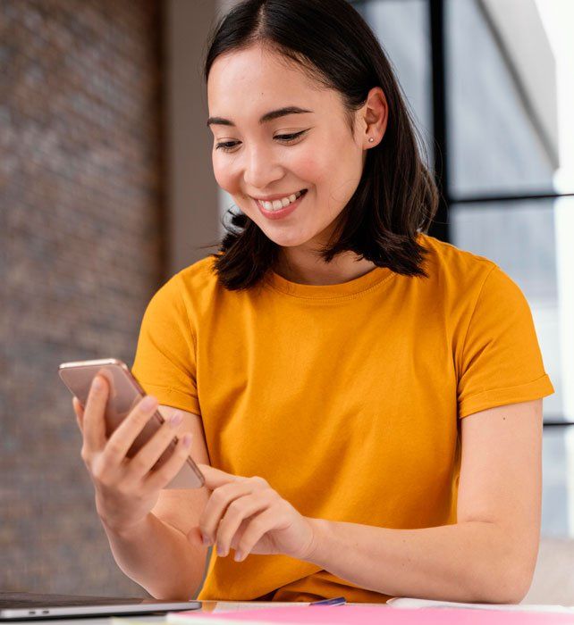A woman in a yellow shirt is smiling while looking at her cell phone.