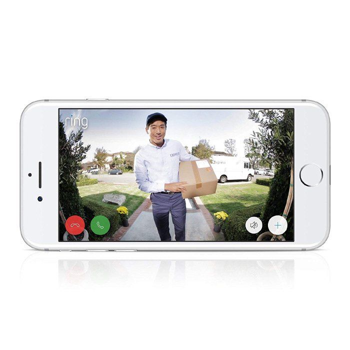 A man is holding a box while standing on a sidewalk in front of a doorbell on a cell phone.