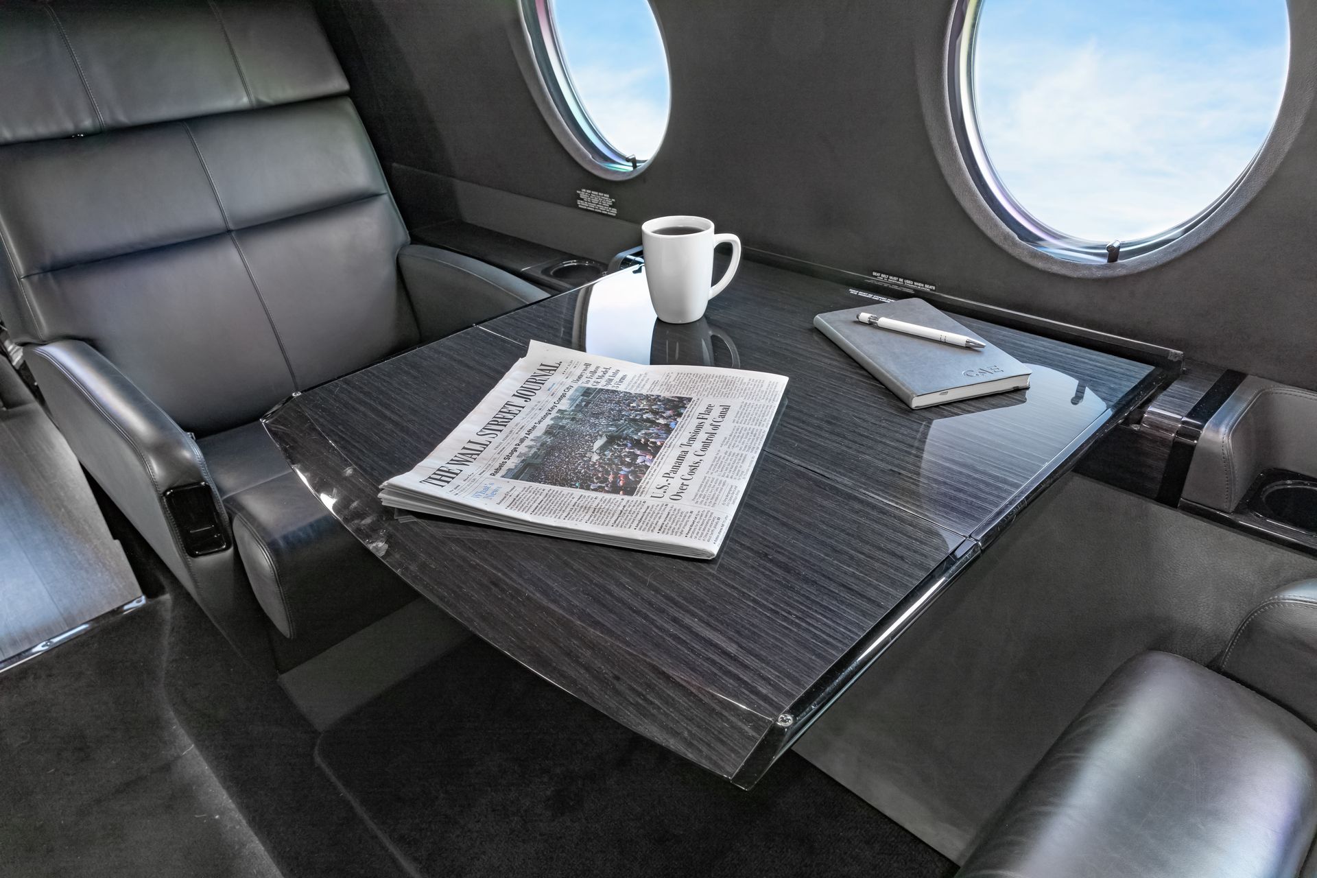 Interior of a private jet with table, coffee, newspaper, and notepad, near oval windows.
