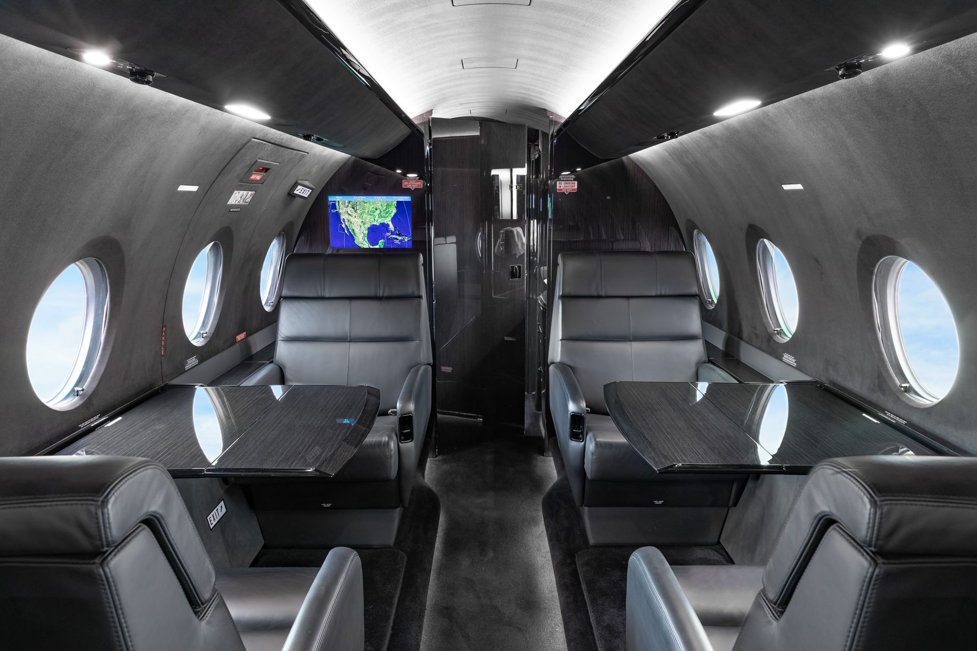 Interior of a private jet with dark gray leather seating, tables, and porthole windows showing blue sky.