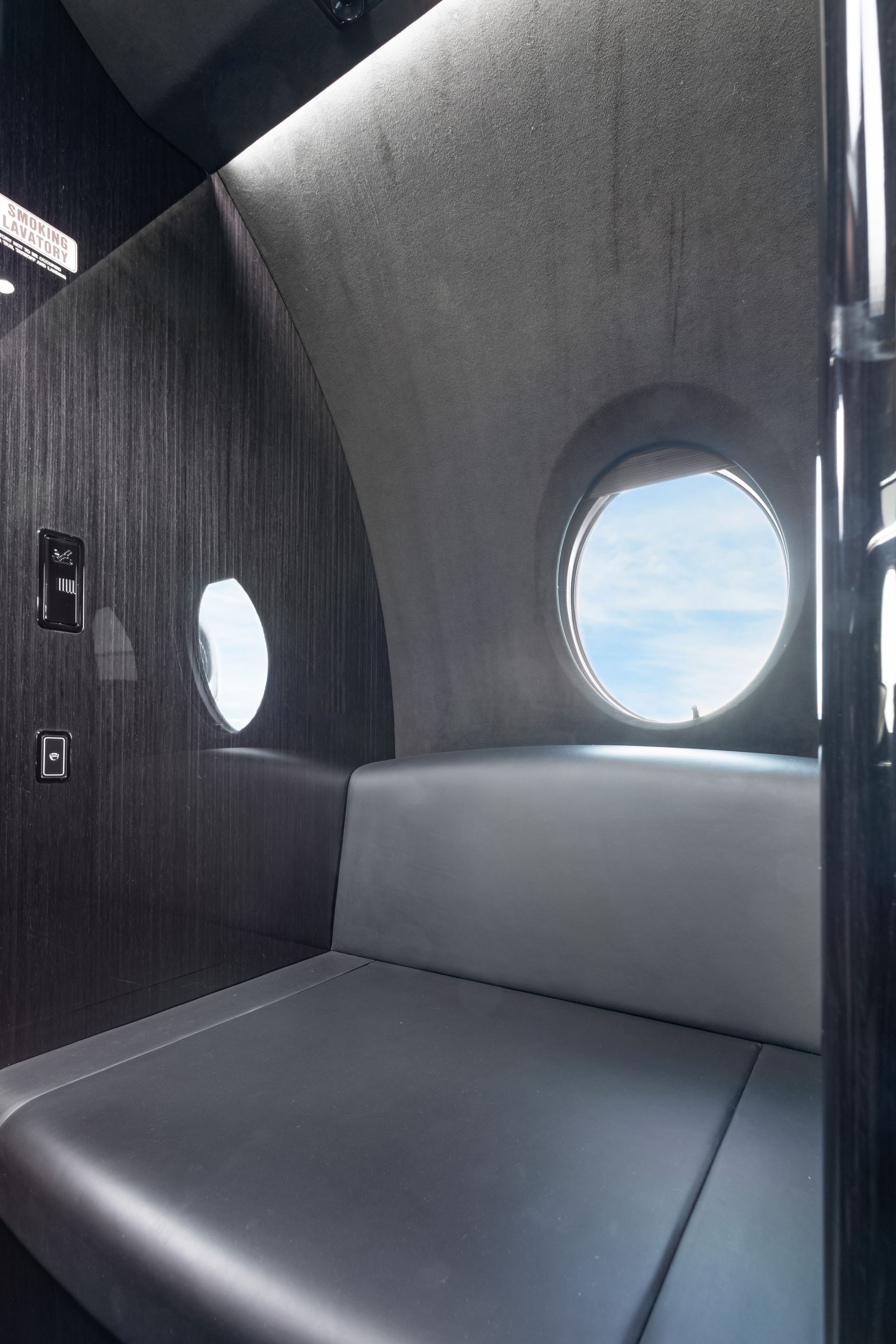 Cozy cabin interior of a private jet with a gray bench seat and porthole window overlooking blue sky.