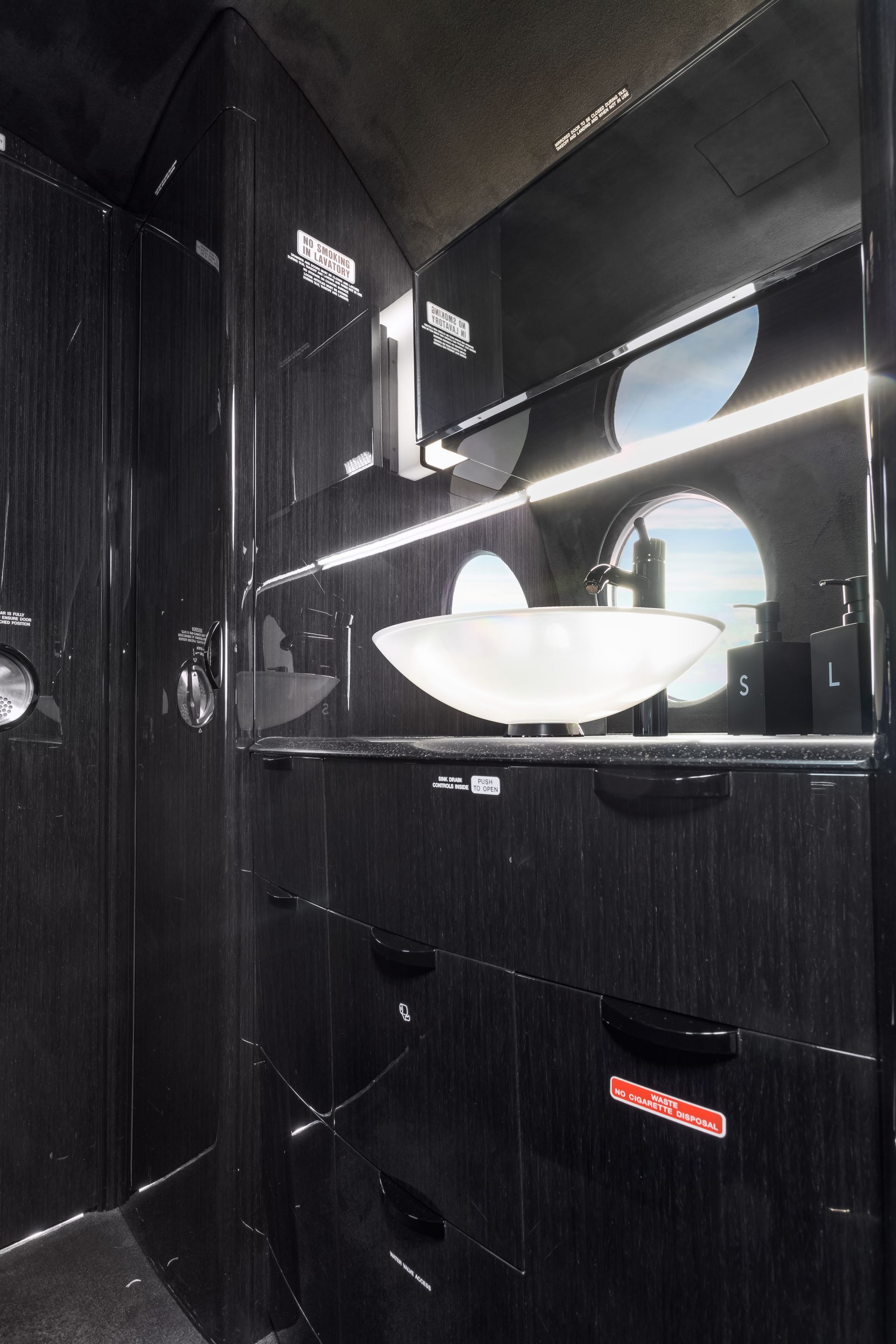 Black airplane bathroom interior with sink, shelving, and porthole windows showing outside view.