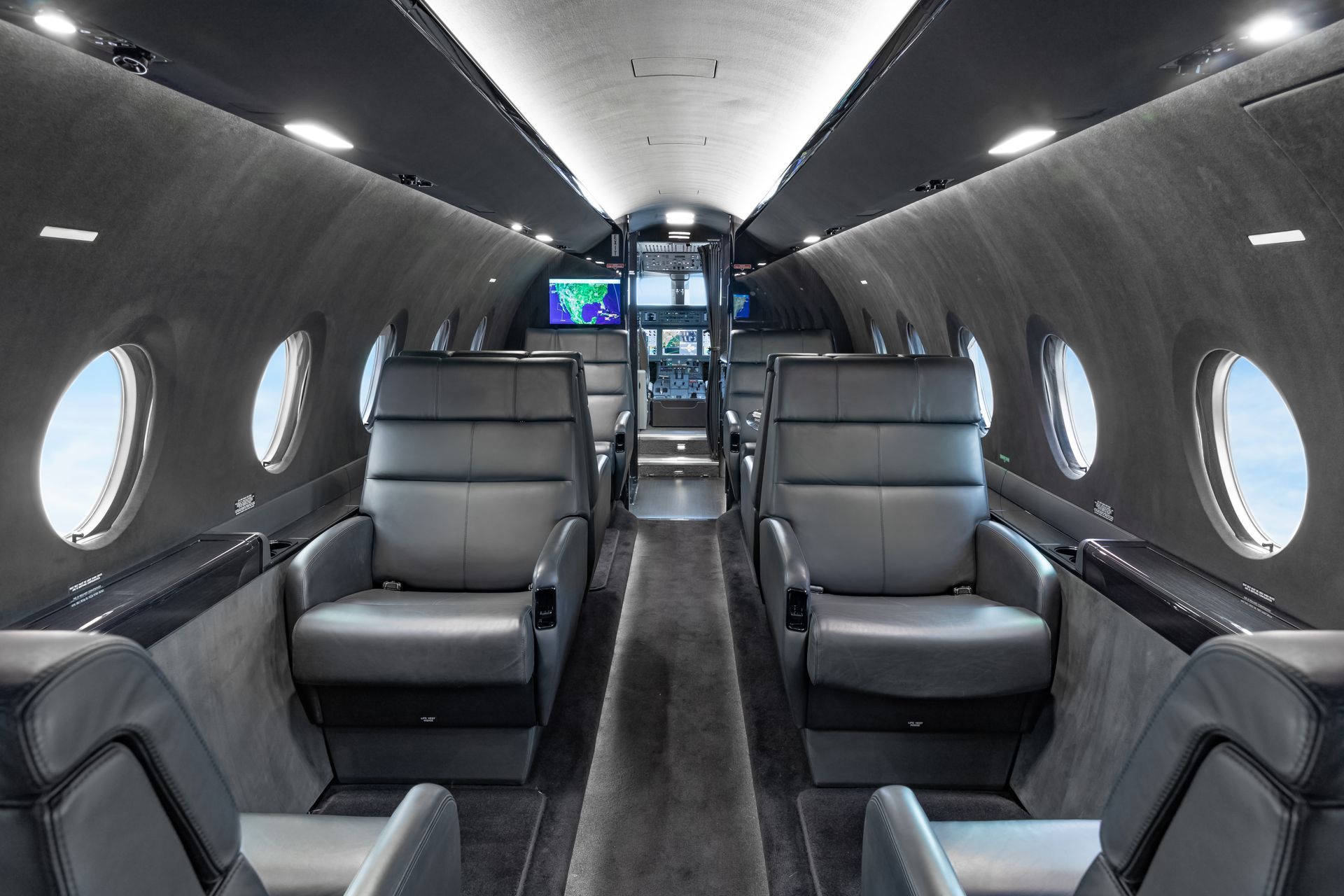 Interior of a private jet with dark gray leather seats, porthole windows, and a carpeted aisle.