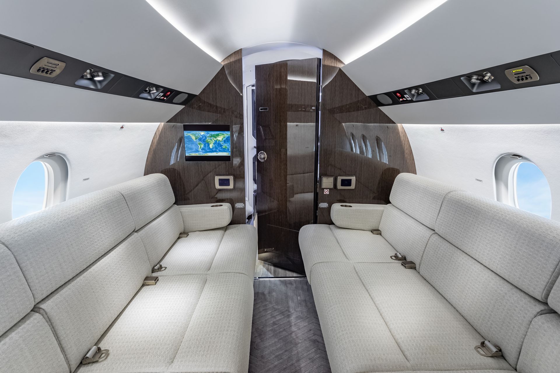 Interior of a private jet cabin, featuring white seating, wood paneling, and a small monitor.
