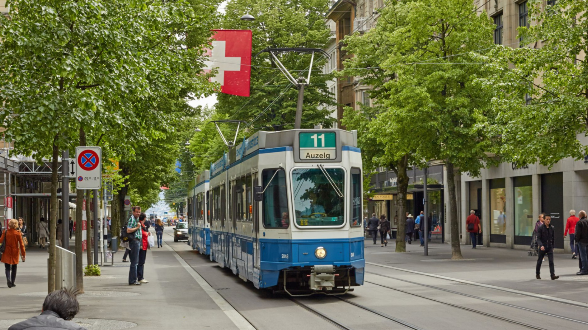 Tram on tracks in Zurich, Switzerland, with Swiss flag above. People walk along the sidewalk.