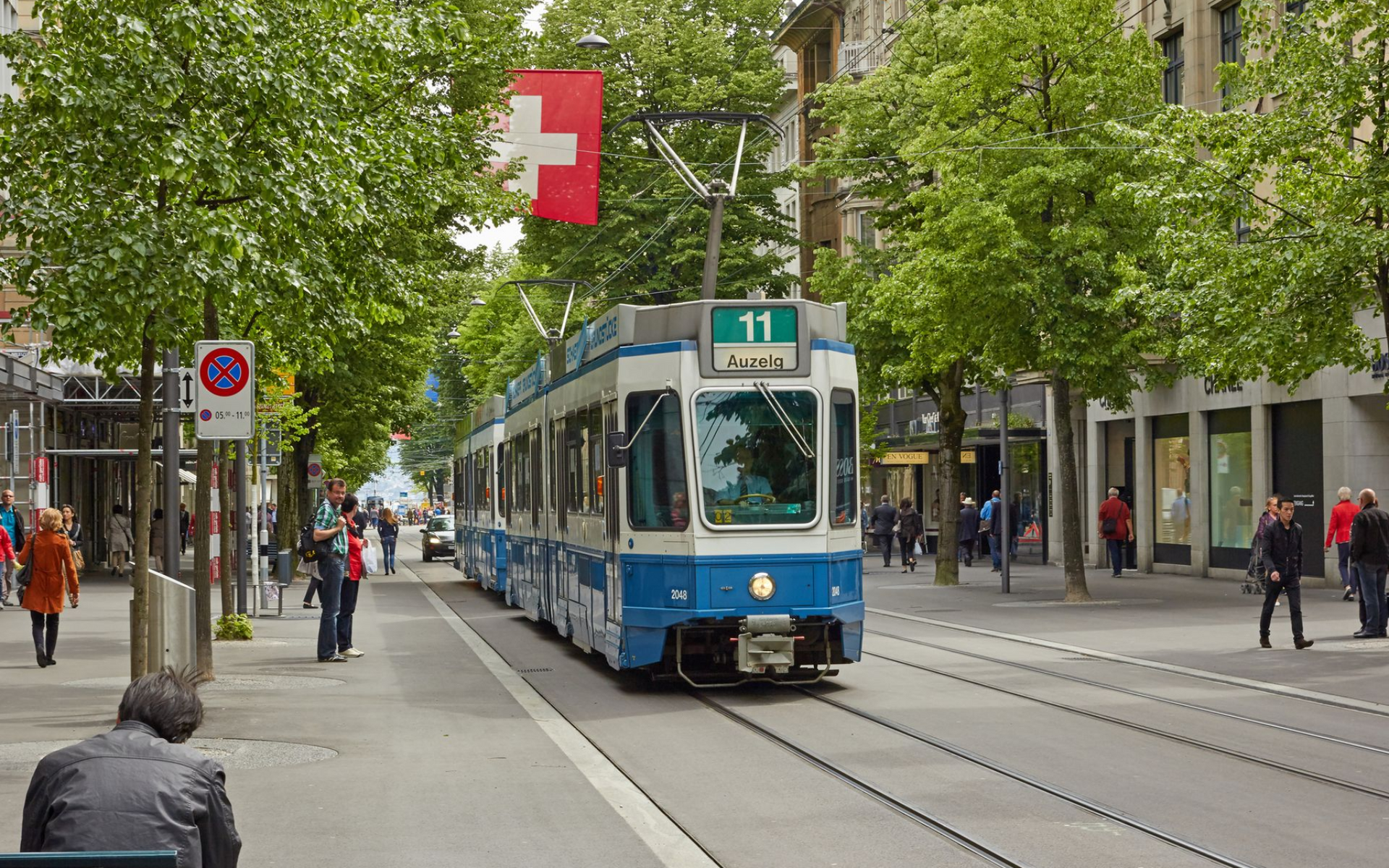 Tram on tracks, white and blue, Swiss flag above. People walk on sidewalks of Bahnhofstrasse lined with trees.