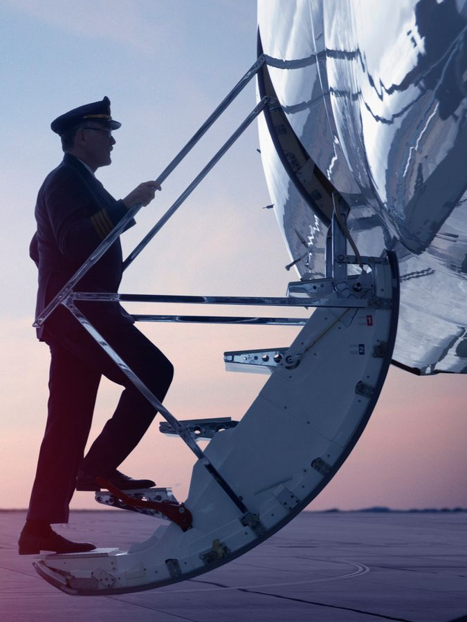 Pilot in uniform climbing stairs to enter airplane on a tarmac at sunset.