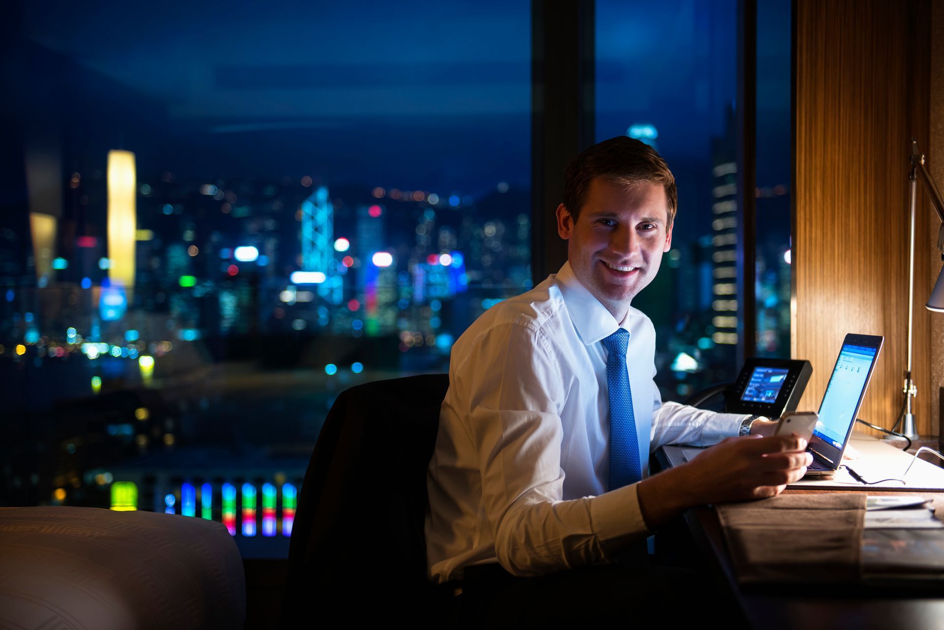Man smiling at a desk, night cityscape in background.