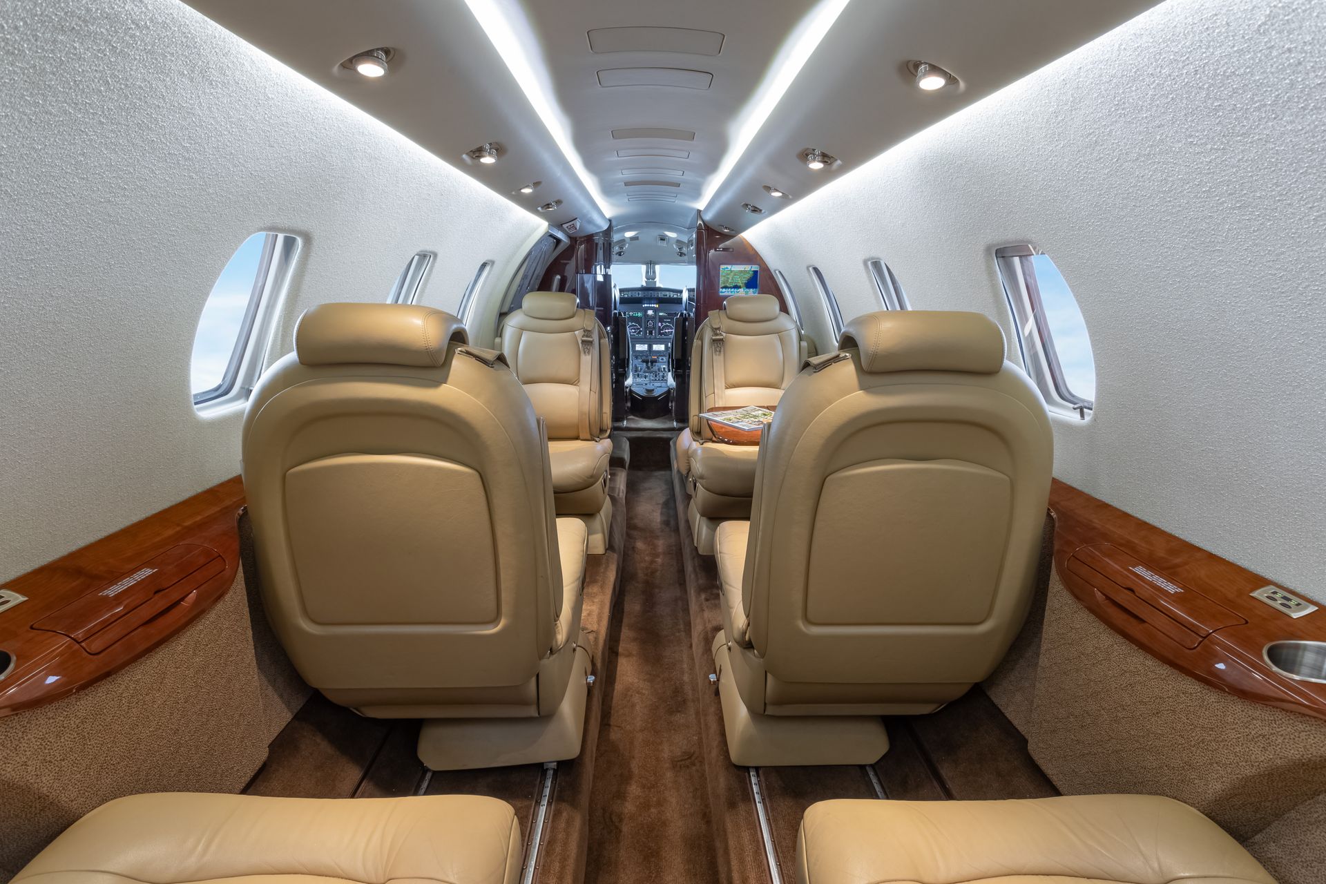 Interior of a private jet cabin, beige leather seats, wood trim, windows, and overhead lighting.