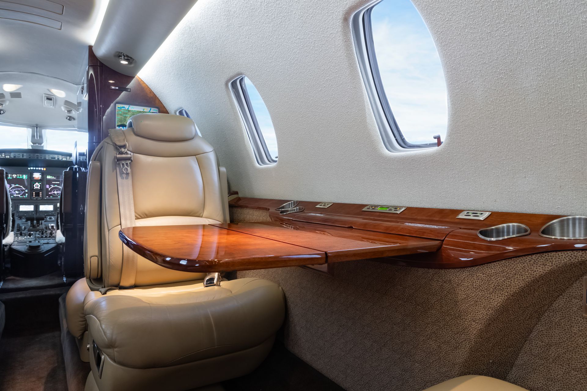 Interior of private jet with beige leather seat, wooden table, and window. Cockpit visible in background.