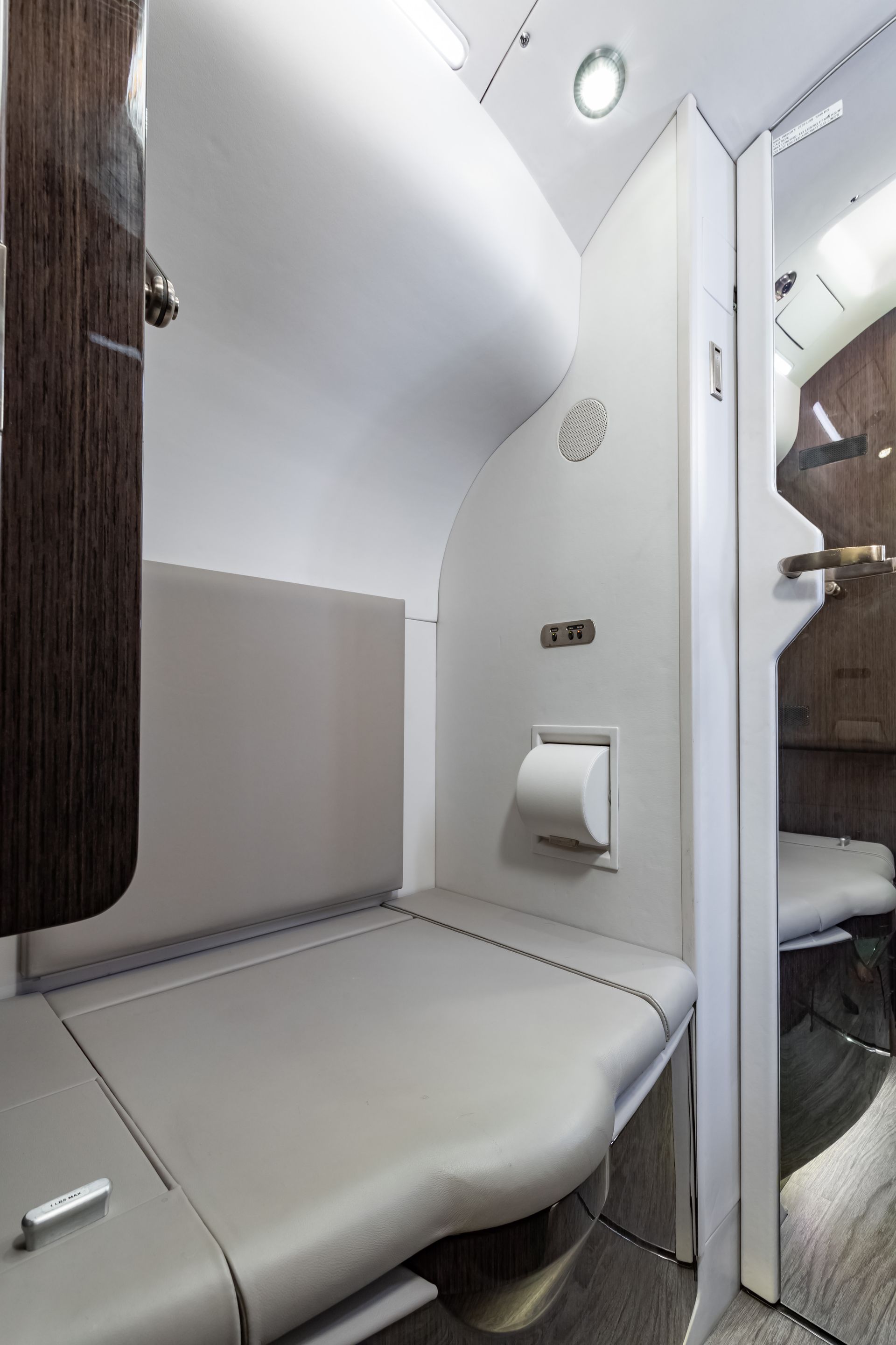 Small, modern aircraft lavatory; cream and wood interior, built-in toilet, gray padded seating.