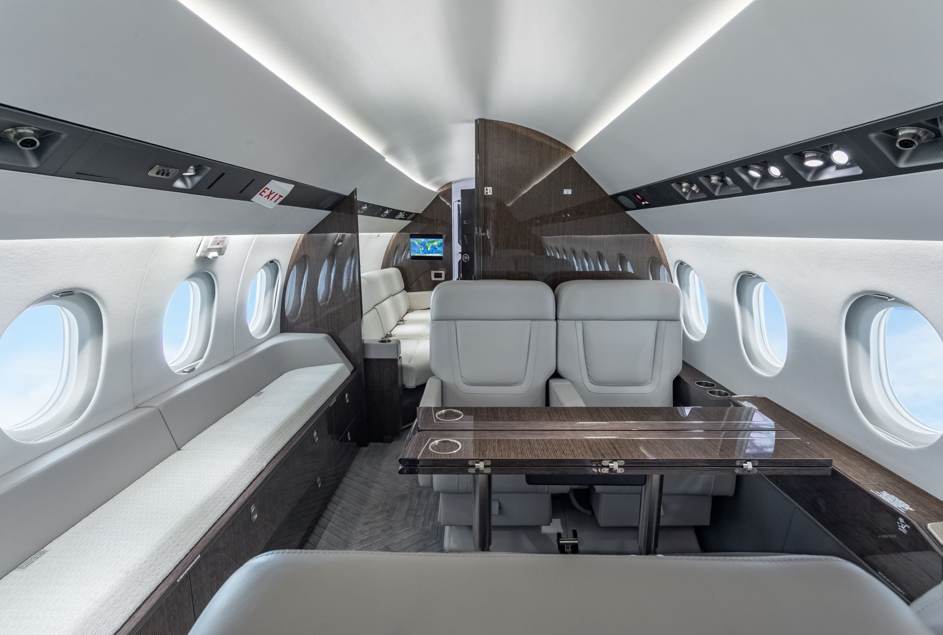 Interior of a luxury private jet with gray seating, dark wood paneling, and oval windows showing the sky.