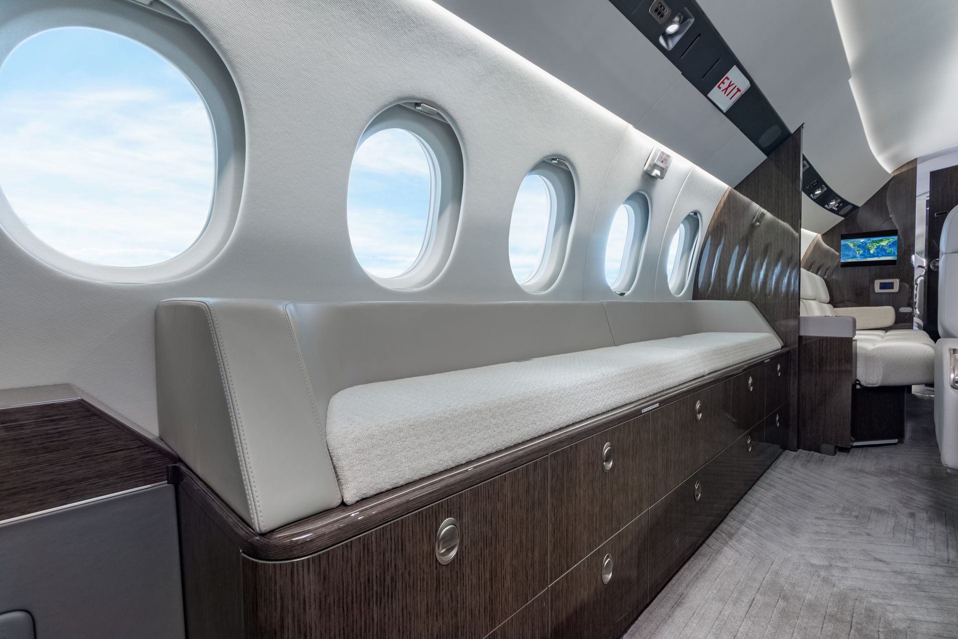 Interior view of a private jet cabin with windows, seating, and wooden cabinetry; sky visible outside.