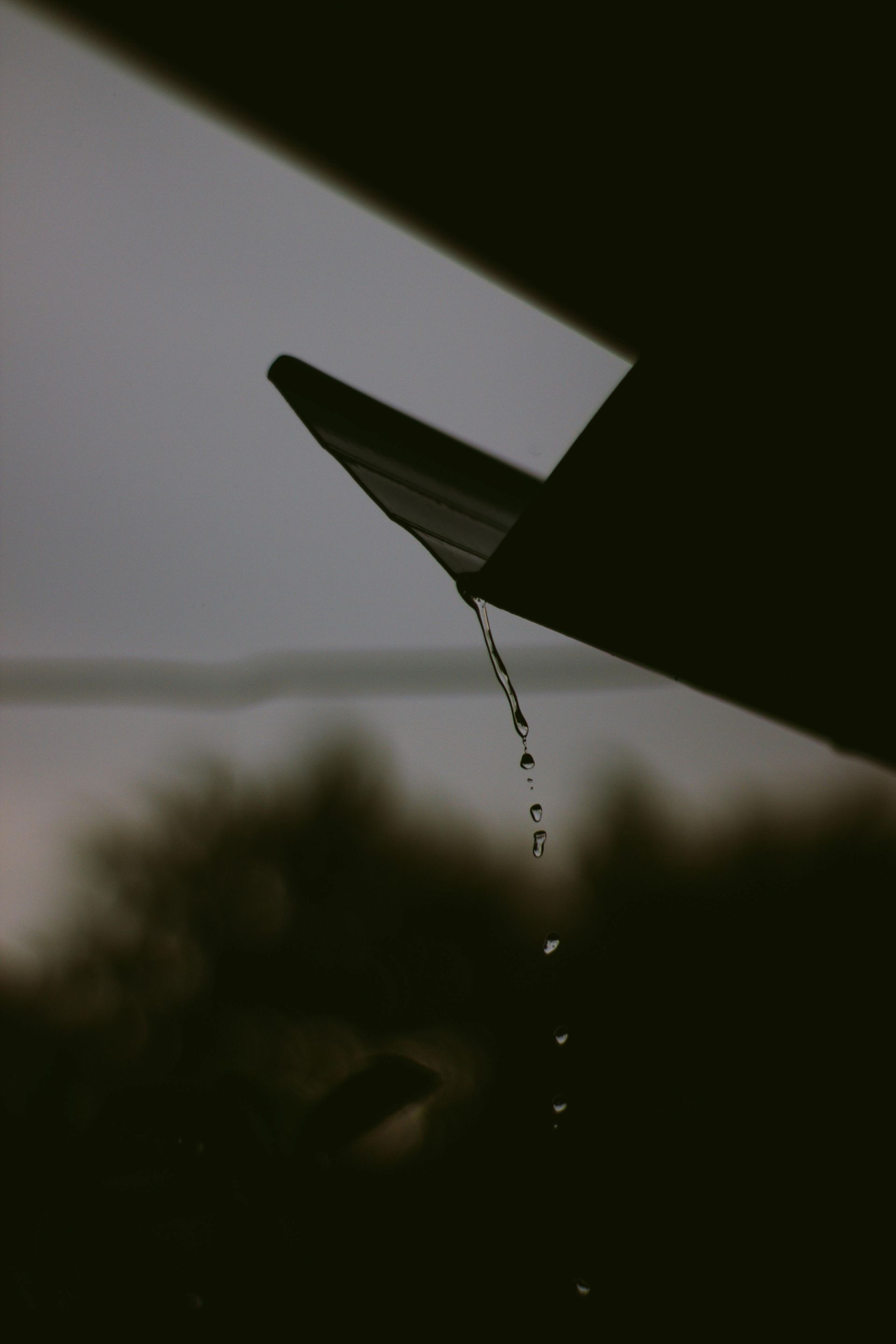 Raindrops dripping from a dark rooftop edge against a blurred, overcast landscape.